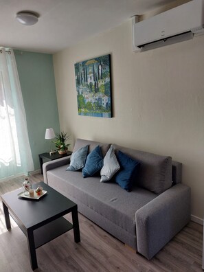 Een woonkamer met een grijze bank, een zwarte salontafel, een schilderij aan de muur en een aan het plafond gemonteerde airconditioner.