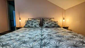 A bedroom with a floral-patterned bedspread, two bedside lamps, and a view of a staircase. - Véro's Lodges. Duplex 5 minutes from Verdun, private parking, garage for two-wheeled vehicles, air conditioning (Thierville-sur-Meuse)