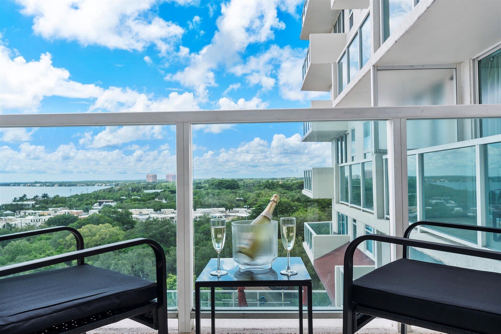 A balcony with a view of a cityscape and a body of water. There is a table with a bottle of champagne and two glasses.