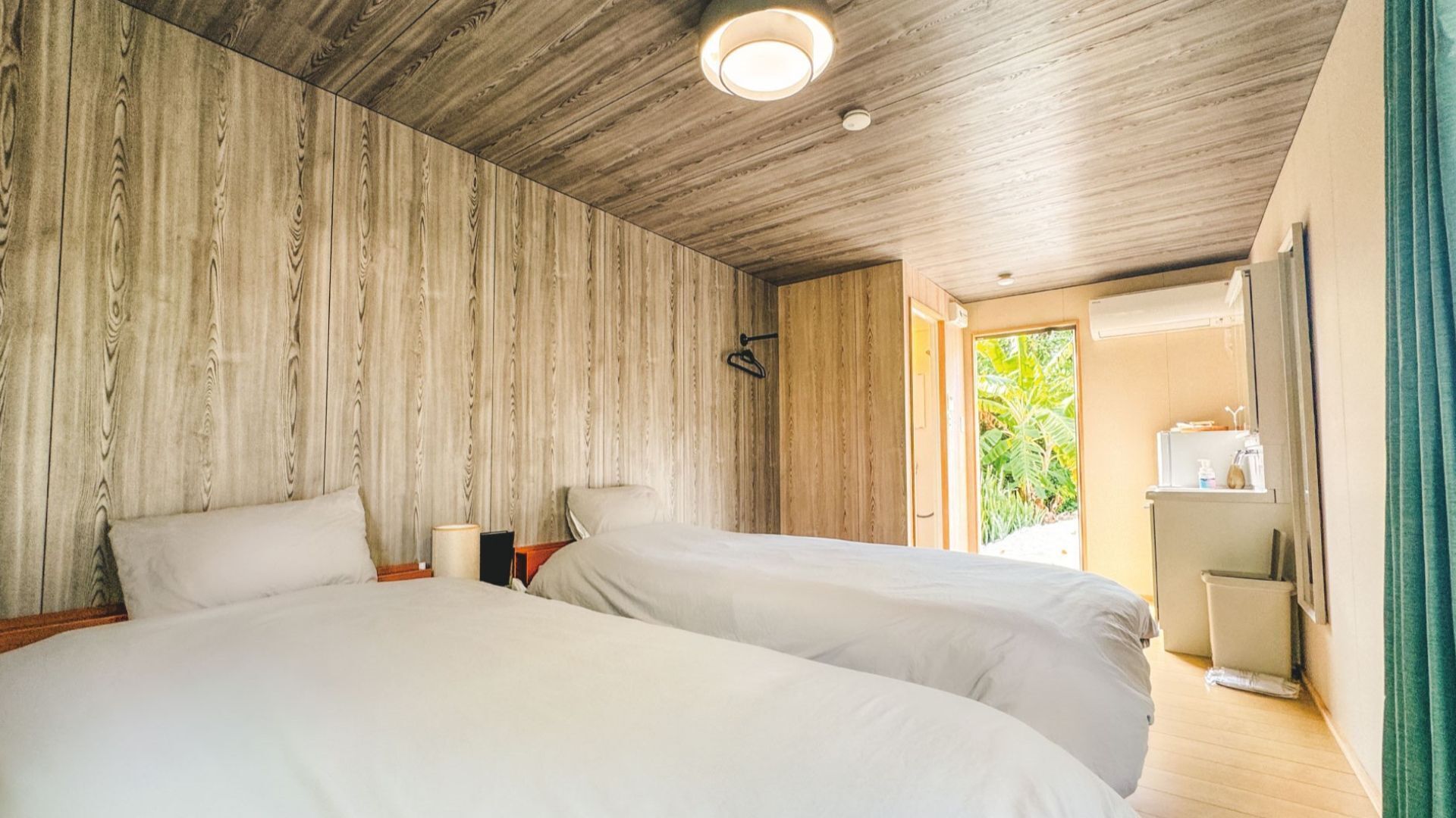 A room with two beds, wooden paneling on the walls, a ceiling light, and a door leading outside.
