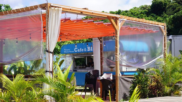 Un pequeño café al aire libre con un letrero azul, mesas y sillas de madera y plantas tropicales.