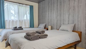 Habitación de hotel con dos camas, cada una con una toalla doblada con cuidado. La habitación tiene un ventanal con cortinas de gasa, un panel de pared de madera y un cabecero de madera.