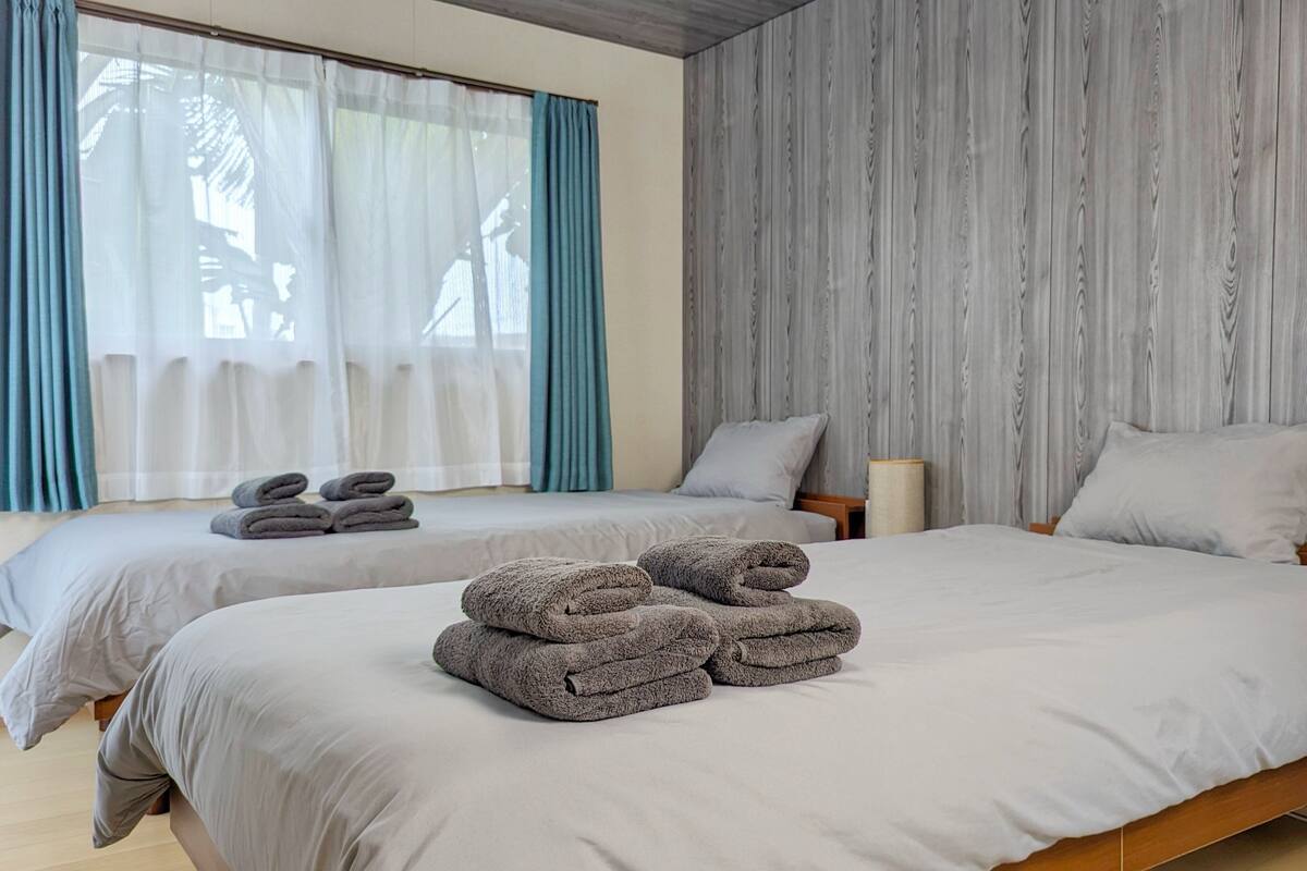 Ein Hotelzimmer mit zwei Betten, jeweils mit einem ordentlich gefalteten Handtuch. Das Zimmer verfügt über ein großes Fenster mit durchsichtigen Vorhängen, eine Holzvertäfelung und einen hölzernen Bettrahmen.