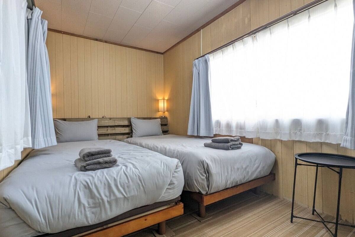 Ein Zimmer mit zwei Betten, einem kleinen Tisch und einem Fenster mit Vorhängen.
