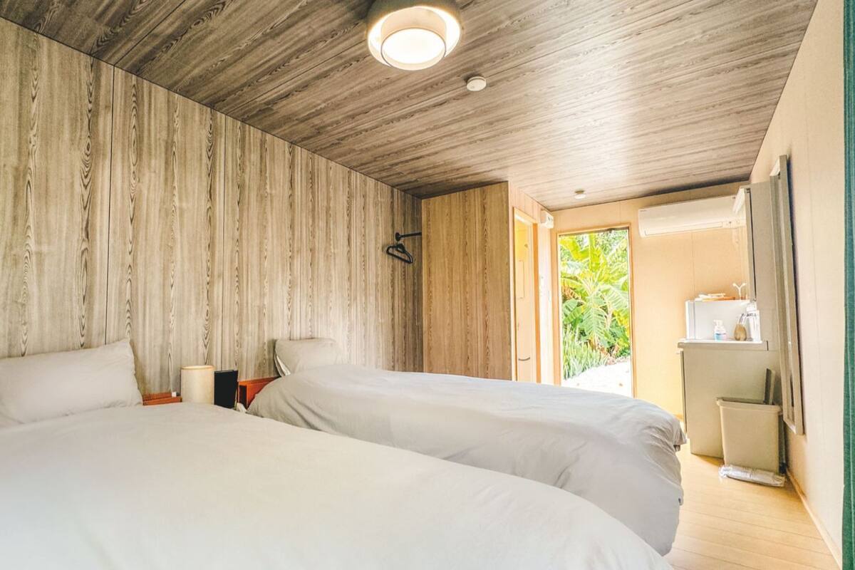 Ein Zimmer mit zwei Betten, Holzwänden, einer Deckenleuchte, einer Tür, die nach draußen führt, und einem kleinen Tisch mit Lampe.