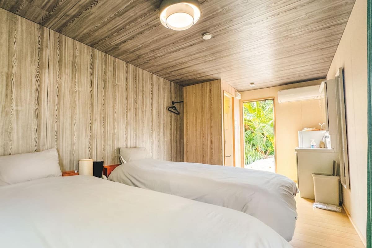 Une pièce avec deux lits, des murs en bois, un luminaire au plafond, une porte donnant sur l’extérieur et une petite table avec une lampe.
