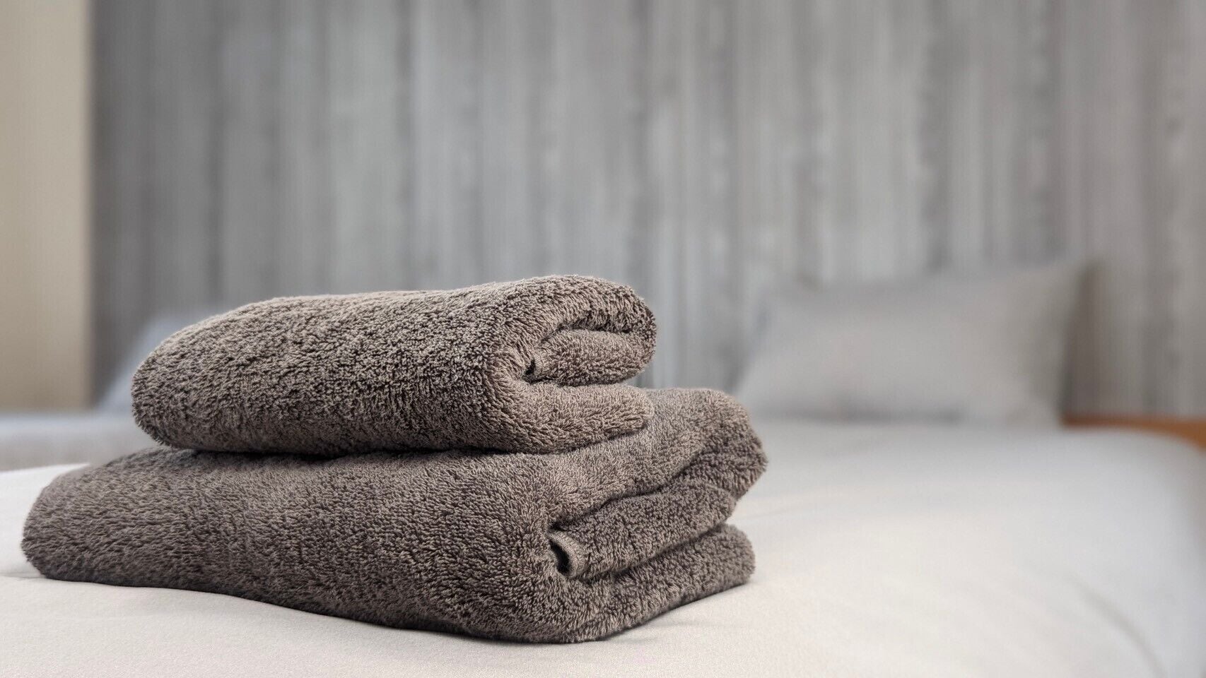Una toalla gris doblada con cuidado sobre una cama con una colcha de color claro y un cabecero gris al fondo.