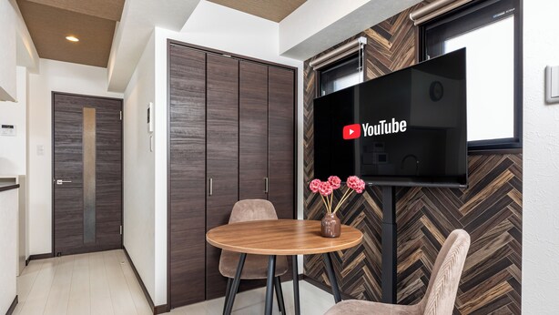 A modern room with a wooden herringbone floor, a round wooden table with two chairs, a flat-screen TV mounted on the wall, and a door with a vertical wooden panel. - SeRENO Shinmachi 801 (Osaka)