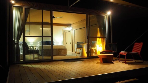 A cozy room with a bed, a small table, and a chair. There is a window with curtains, and a lamp is on. - King tent 5 Nelly bay (Nelly Bay)