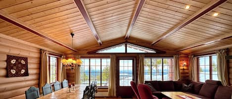 A log cabin interior with a dining area, a living room with a sofa and table, and large windows offering a view.