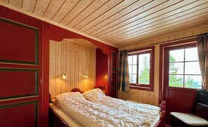 A room with a wooden ceiling, a bed with white bedding, a window with curtains, and a chair.