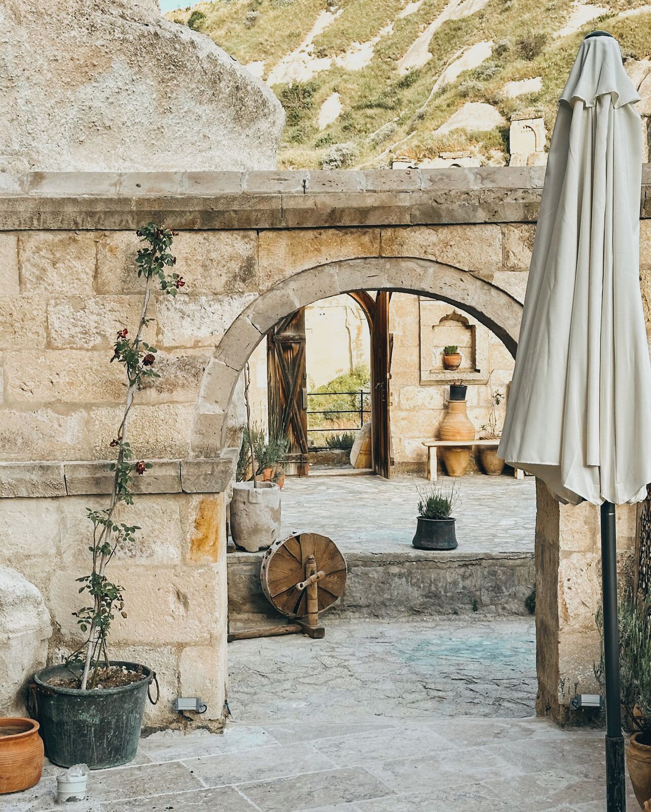 An outdoor courtyard with stone walls, a wooden barrel, potted plants, and an umbrella.