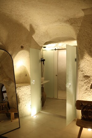Une pièce aux murs de pierre, avec un miroir, un banc en bois et une porte donnant sur une salle de bain équipée d’un lavabo et d’un sèche-cheveux.