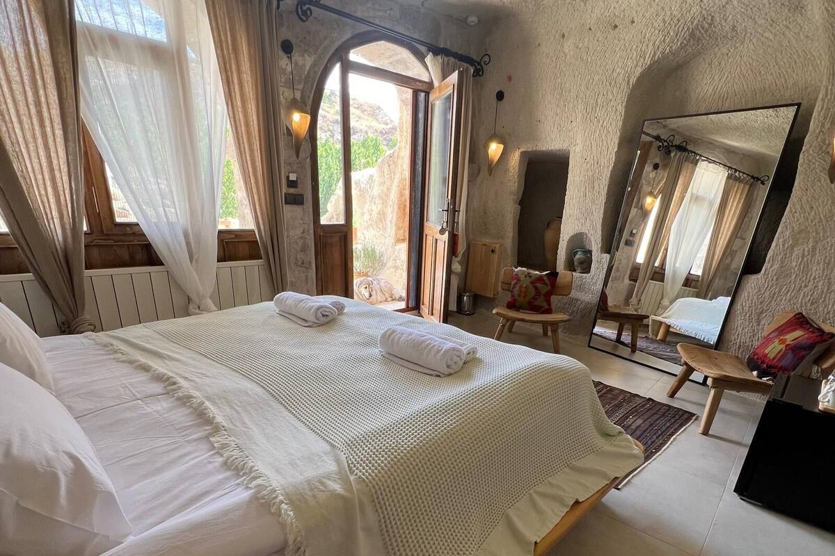 Une chambre avec un grand lit, un banc en bois, une chaise, un miroir et une vue sur l’extérieur.