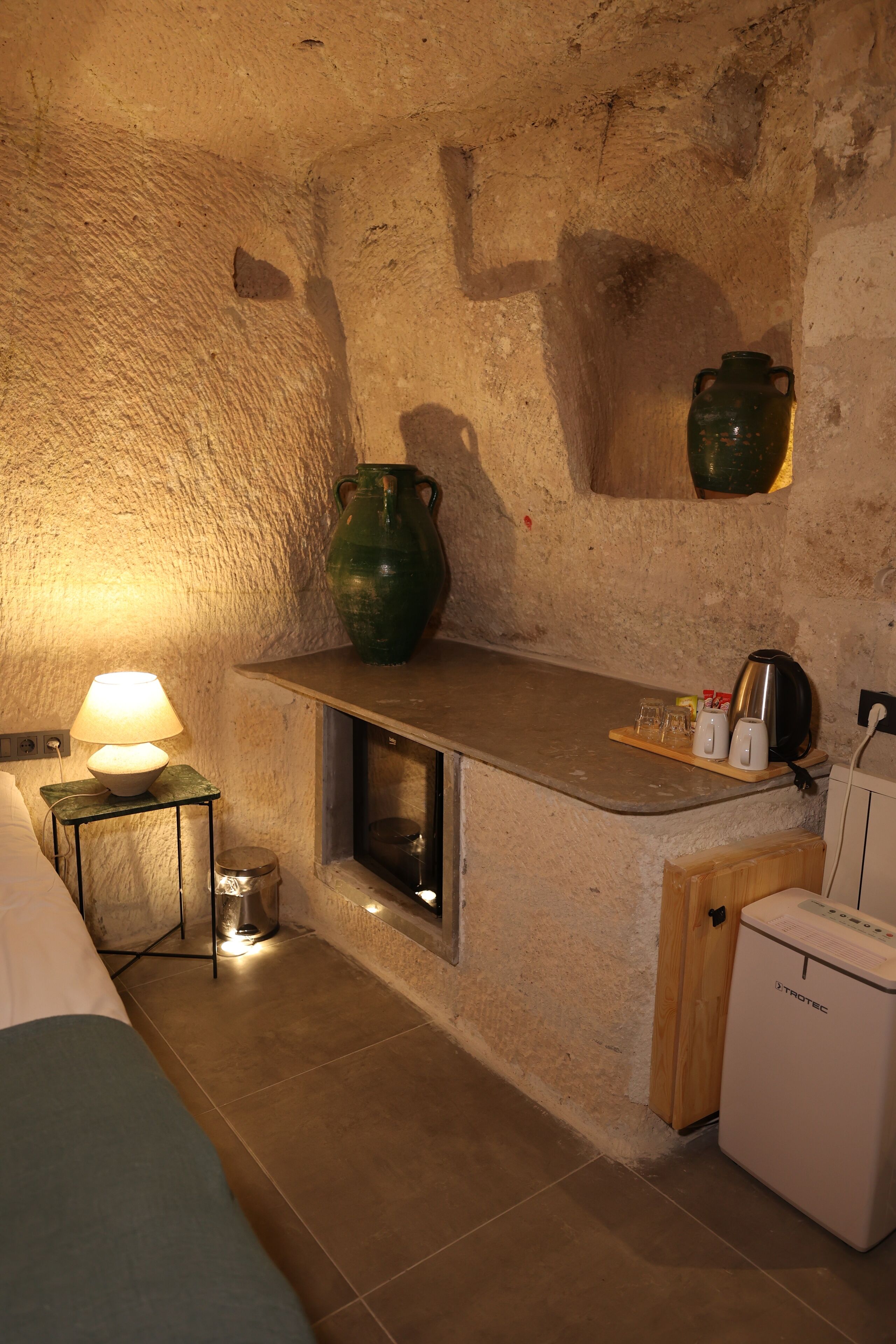Une cuisine rustique avec un mur en pierre, une étagère en bois ornée d’un vase vert, une petite table avec une lampe et un réfrigérateur.