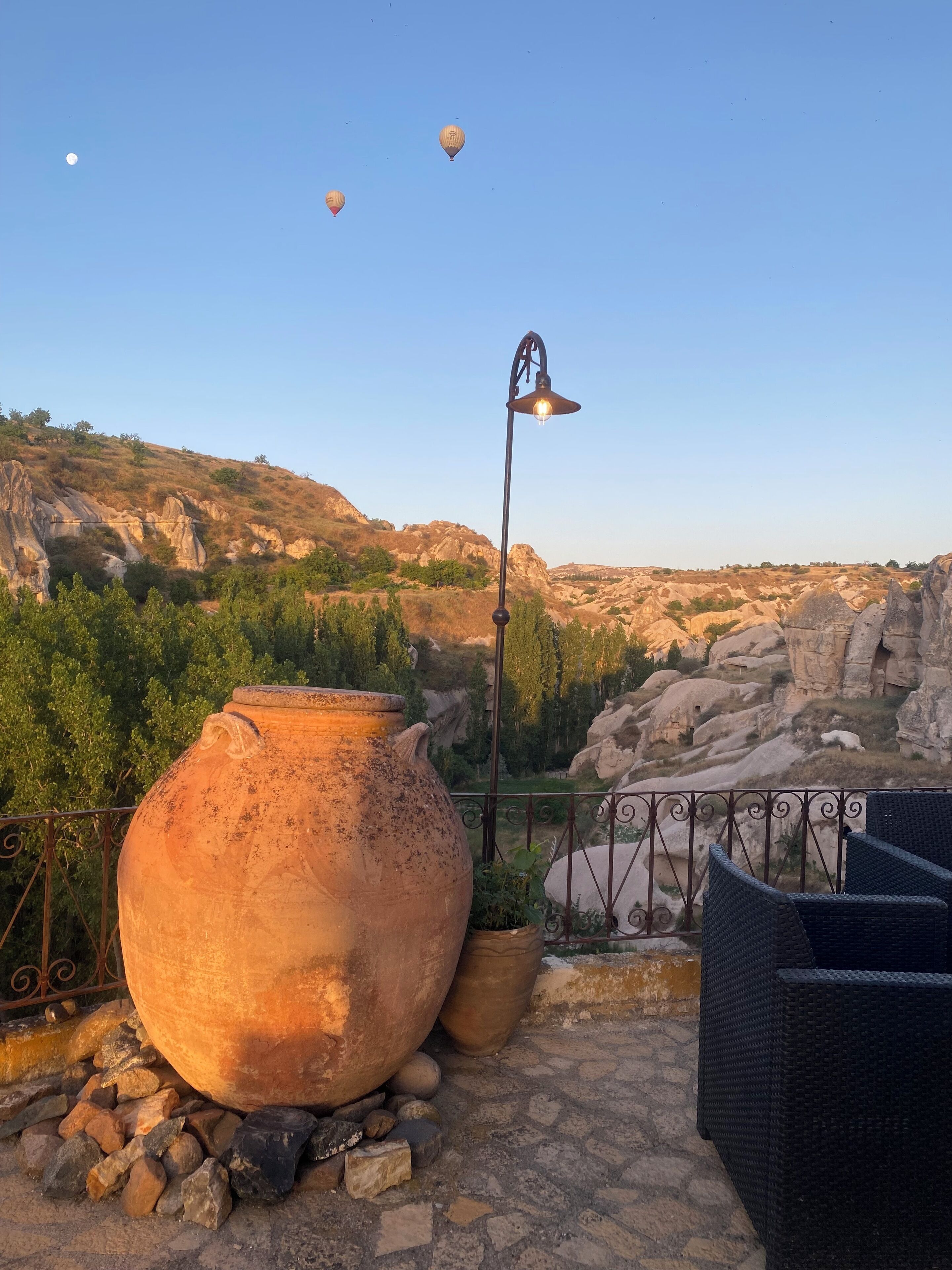 Eine Terrasse mit einem großen Terrakottatopf, Korbmöbeln und einem Metallzaun mit Blick auf felsiges Gelände und einen klaren Himmel.