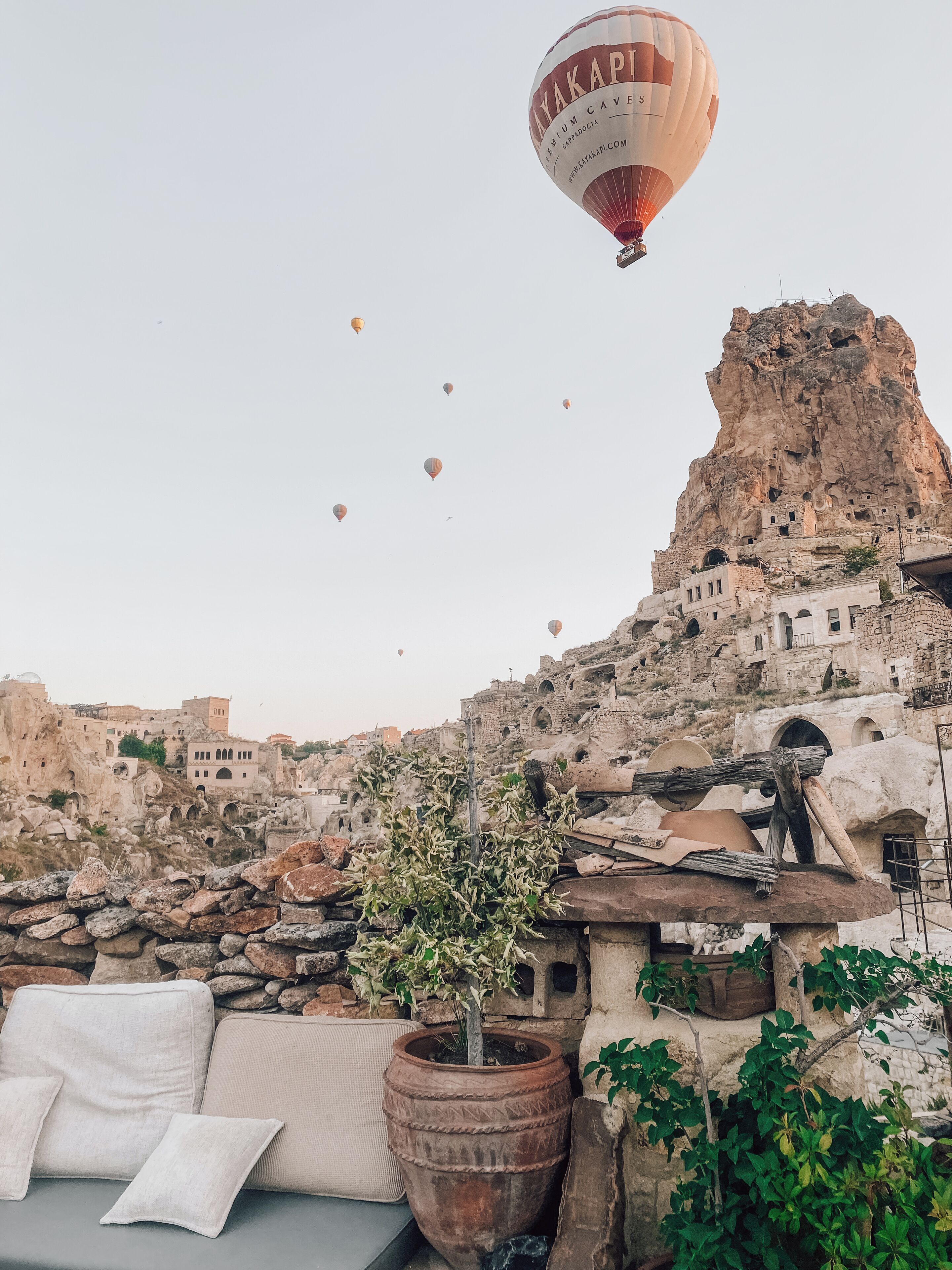 A hot air balloon with "Premium Caves" written on it flying over a rocky landscape with traditional stone buildings.