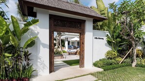 A white building with a wooden archway entrance, a sign reading "Villa Marang," and a view of a pool area with lounge chairs and umbrellas.