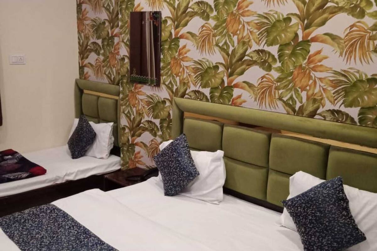 A hotel room with two beds, each with a green headboard and white bedding. A tropical leaf-patterned wallpaper decorates the wall behind the beds.