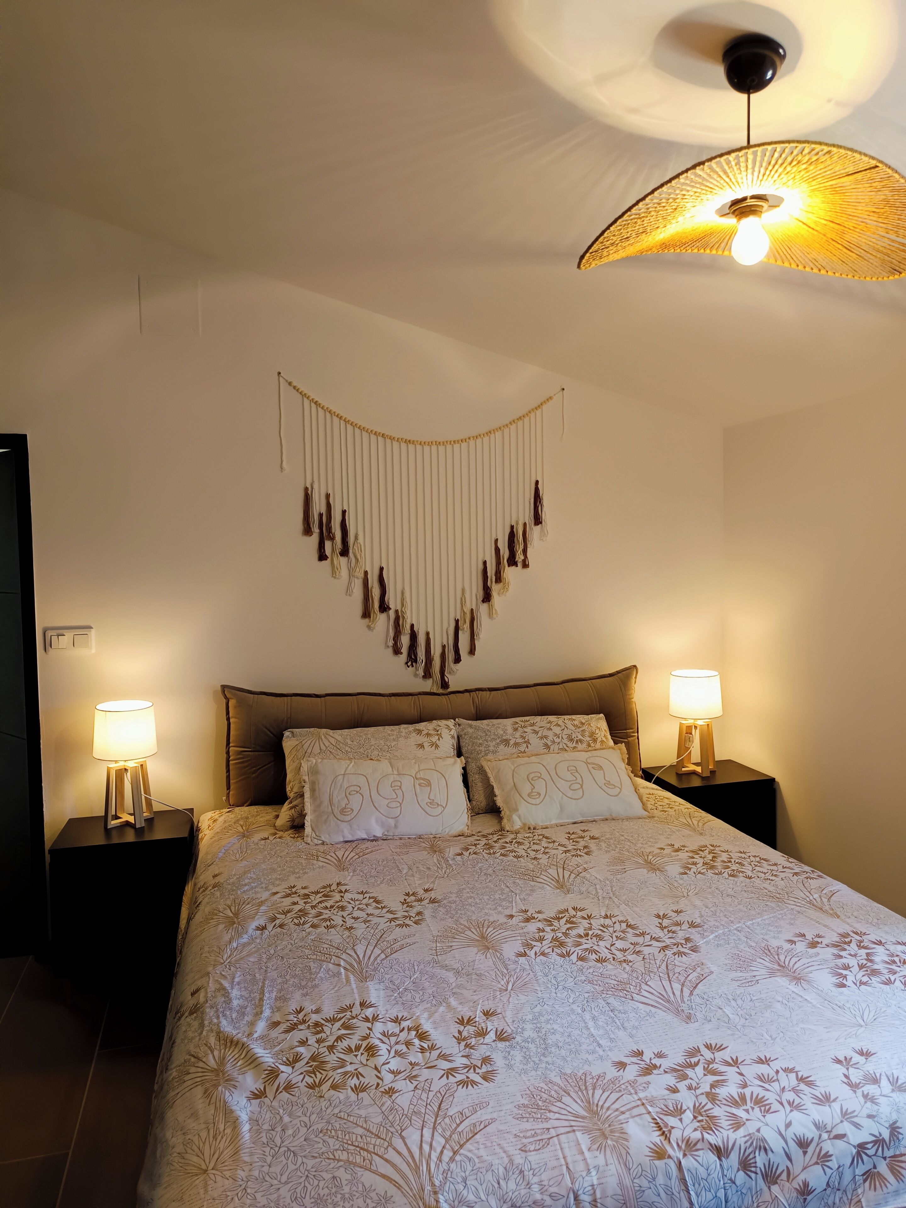 Un dormitorio con cama, dos mesitas de noche con lámparas, un adorno colgante de macramé y un ventilador de techo.