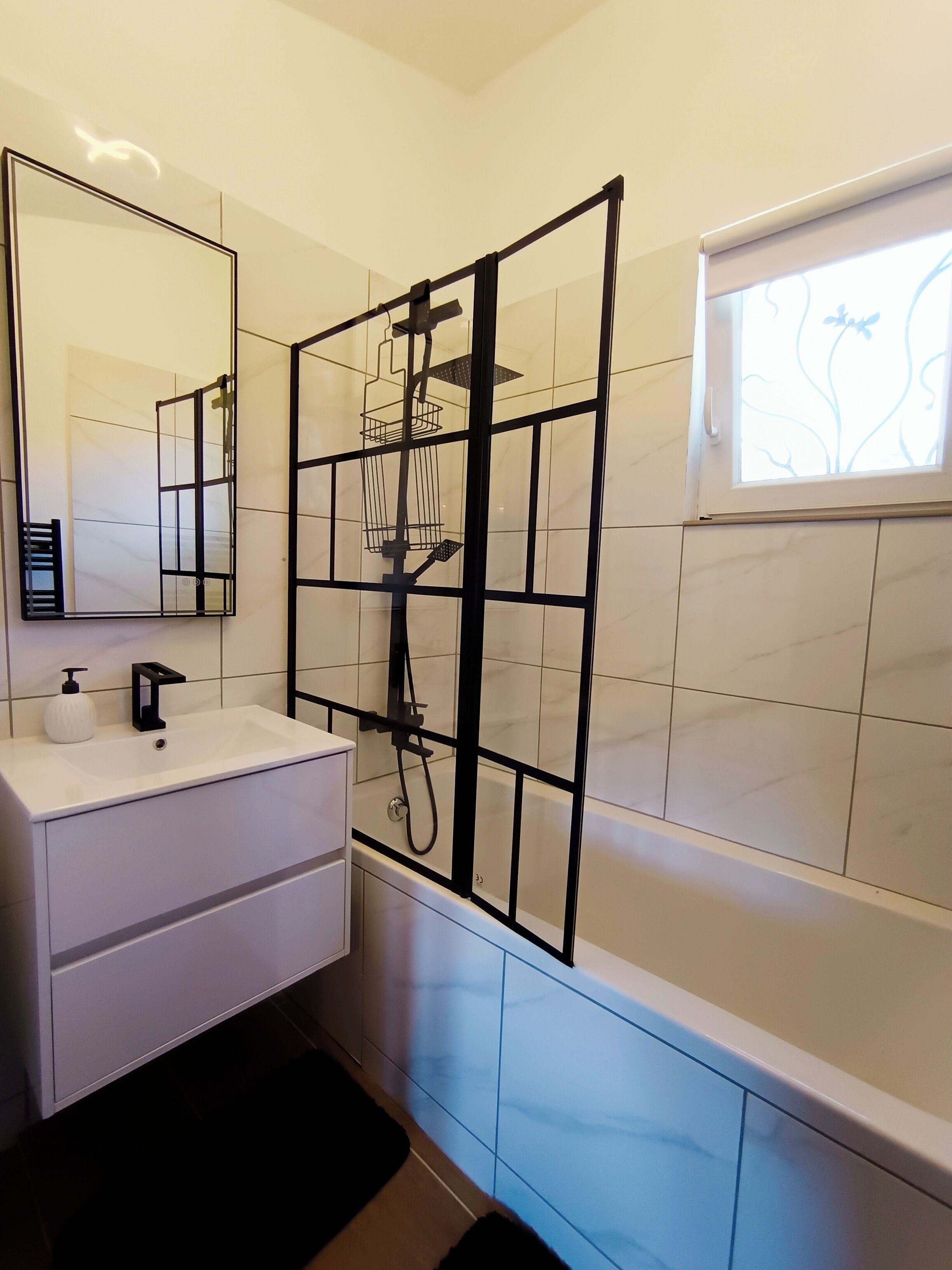 Un baño moderno con tocador blanco, un espejo grande, cabina de ducha y ventana con vista decorativa.
