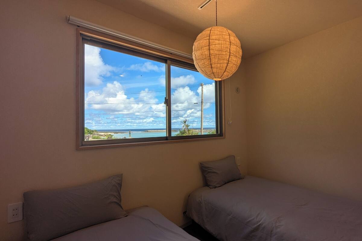 A room with two beds, a large window with a view of the sea, and a hanging light fixture.