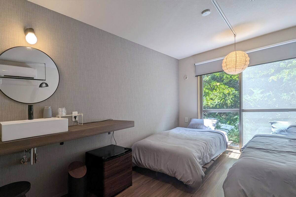 A modern bedroom with a large window, a round mirror, a bedside table, a small cabinet, and a hanging light fixture.