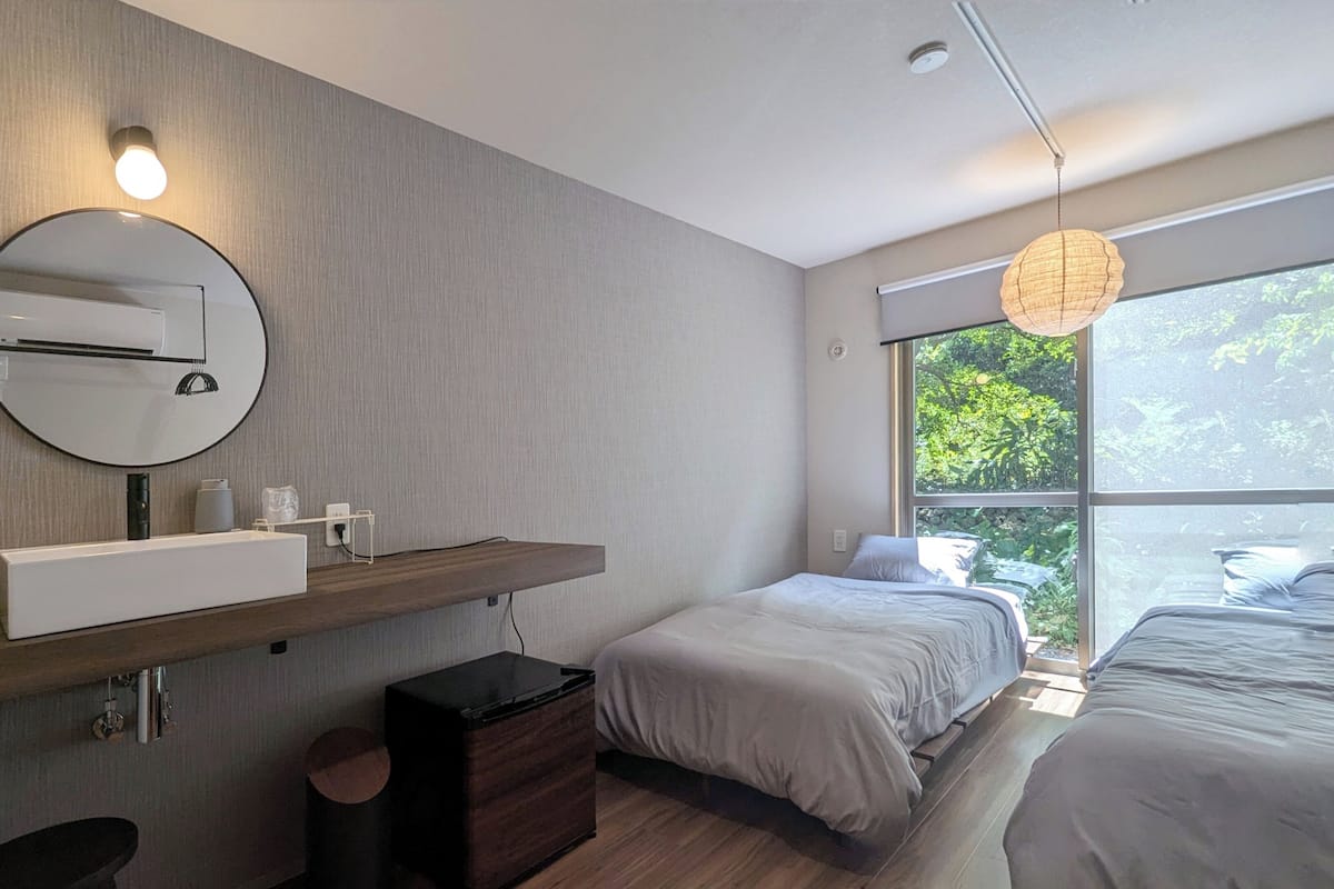 A modern bedroom with a built-in sink, a round mirror, a bedside table, a small cabinet, a hanging light fixture, and a large window with a view of greenery.