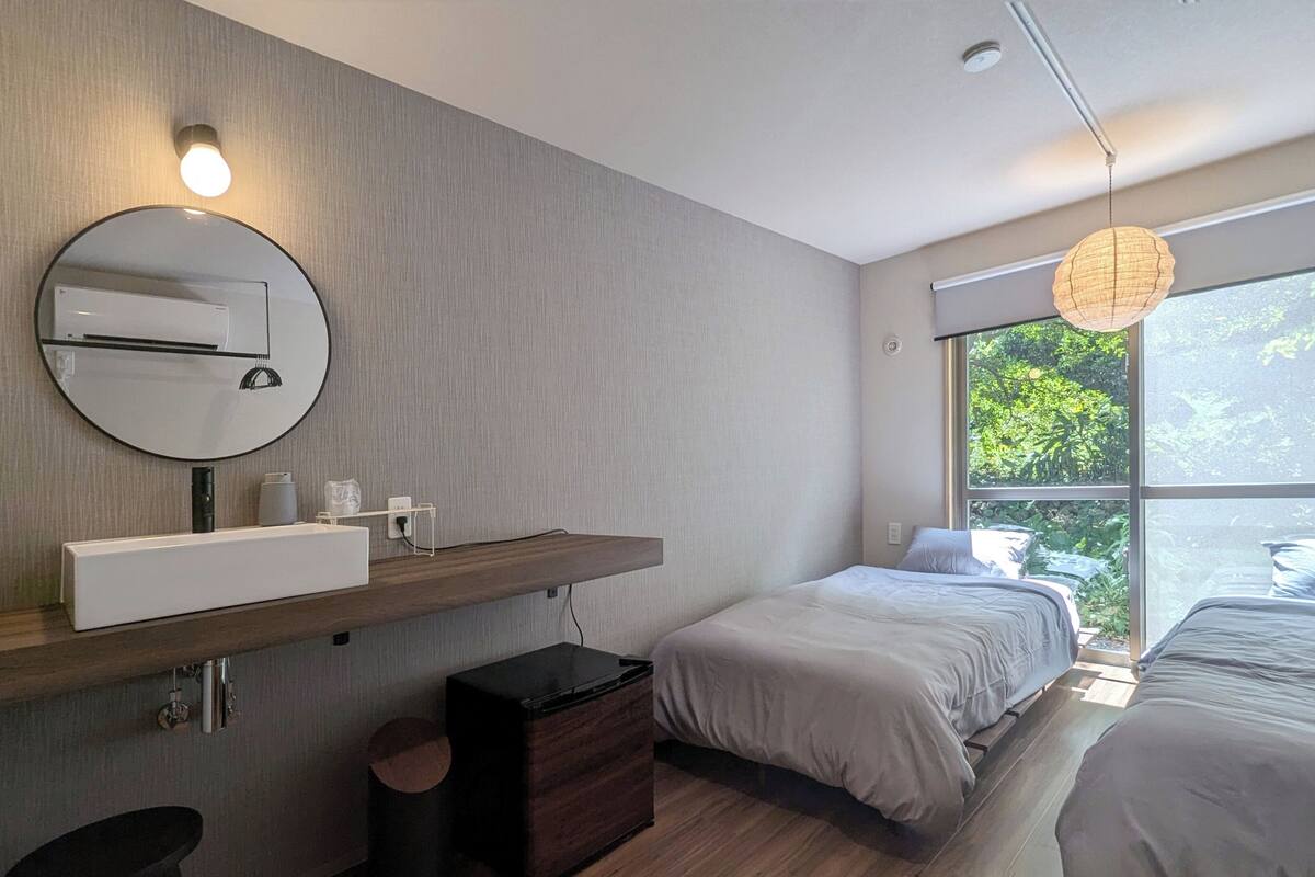 A modern bedroom with a large window, a round mirror, a bedside table, a small cabinet, and a hanging light fixture.