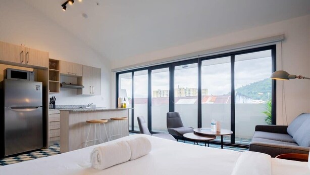 Habitación de hotel con cama, kitchenette, mesa de comedor y balcón con vista a edificios.
