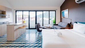 Habitación de hotel moderna con una cama grande, kitchenette, comedor y balcón con vista a la ciudad.