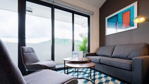 Una habitación moderna con un sofá gris, dos sillones, una mesita redonda con una planta y botellas de agua, y un ventanal con vistas a una colina.