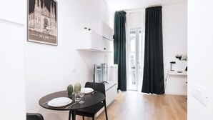 Une pièce moderne et bien éclairée, avec une table à manger dressée pour deux personnes, un téléviseur à écran plat et une grande fenêtre dotée de rideaux foncés.