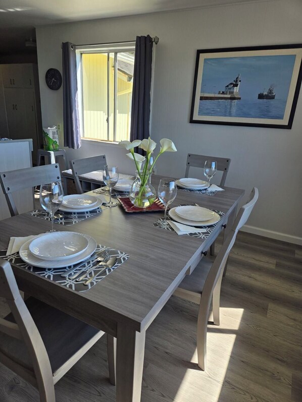 Un comedor con una mesa de madera dispuesta para cuatro personas, sillas, un florero con flores y un cuadro de un barco en la pared.