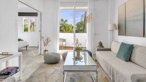 Un salon moderne avec une table basse en verre, un canapé et une table à manger. De grandes fenêtres offrent une vue sur les palmiers.