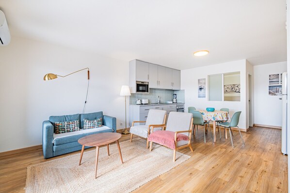 A modern living room with a sofa, coffee table, and chairs, a kitchen area with cabinets and appliances, and a dining table with chairs.