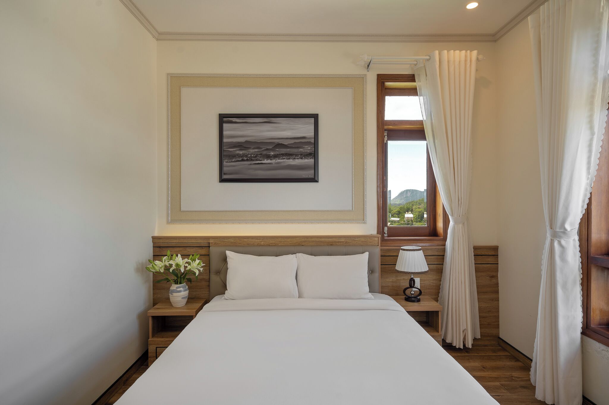 A neatly arranged bedroom with a large bed, a bedside table with a lamp, a nightstand with a vase of flowers, and a framed picture on the wall.