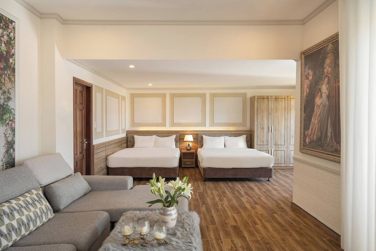 A hotel room with two beds, a sofa, a small table with a vase of flowers, and a painting on the wall.