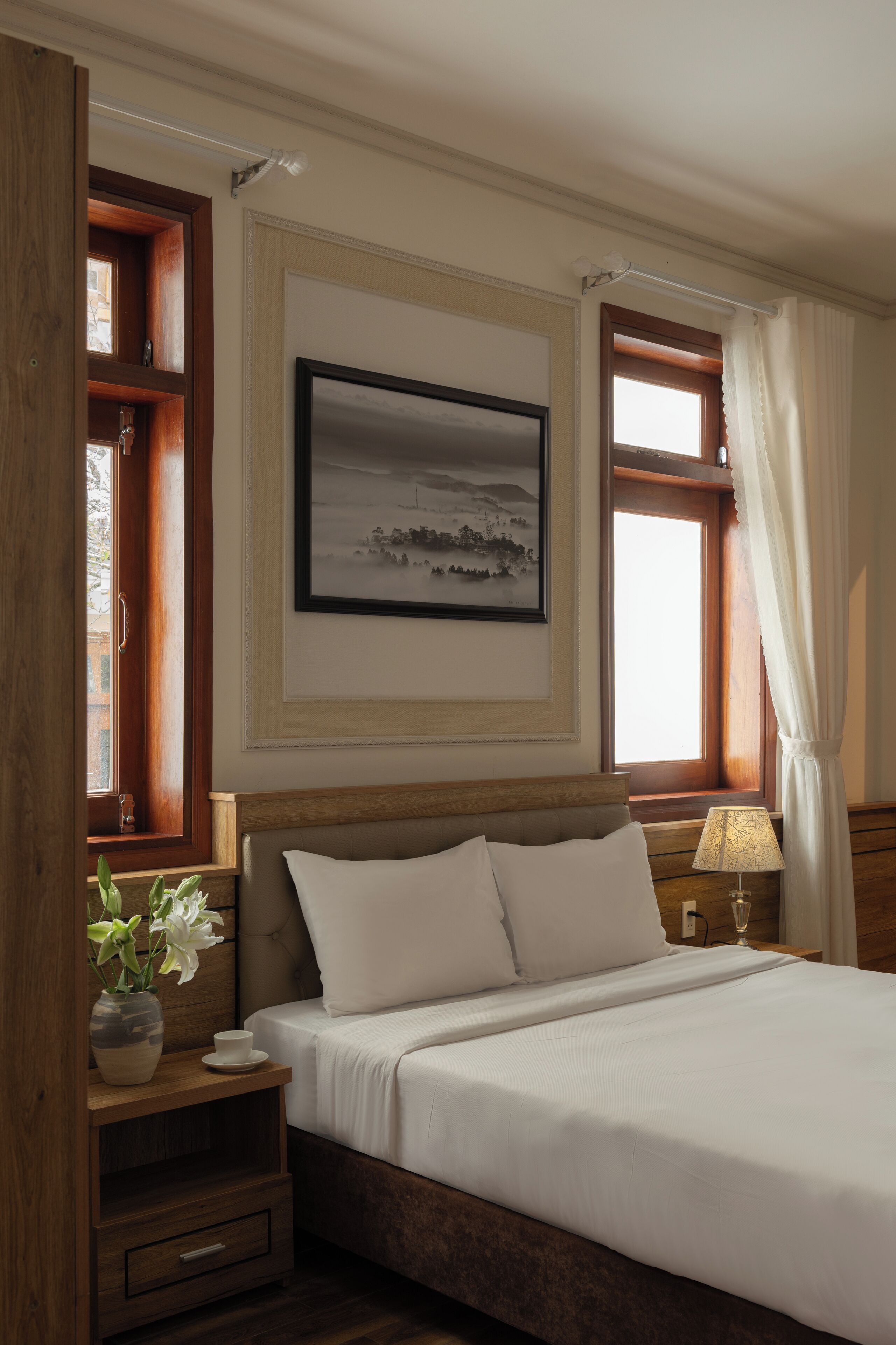 A bedroom with a bed, a bedside table with a lamp, a window with curtains, and a framed picture on the wall.
