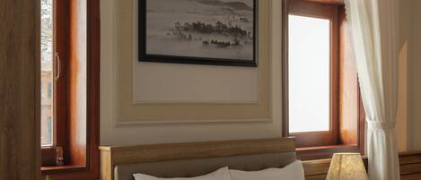 A bedroom with a bed, a bedside table with a lamp, a window with curtains, and a framed picture on the wall.