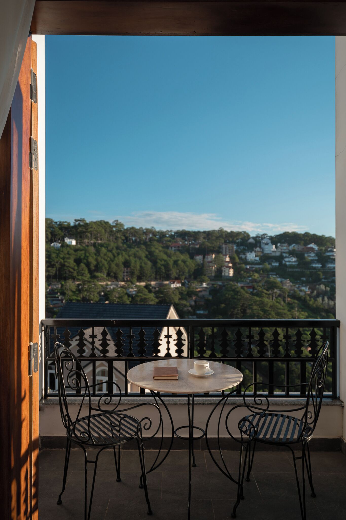 A balcony with a round table and two chairs, overlooking a scenic view of a town with buildings and trees.