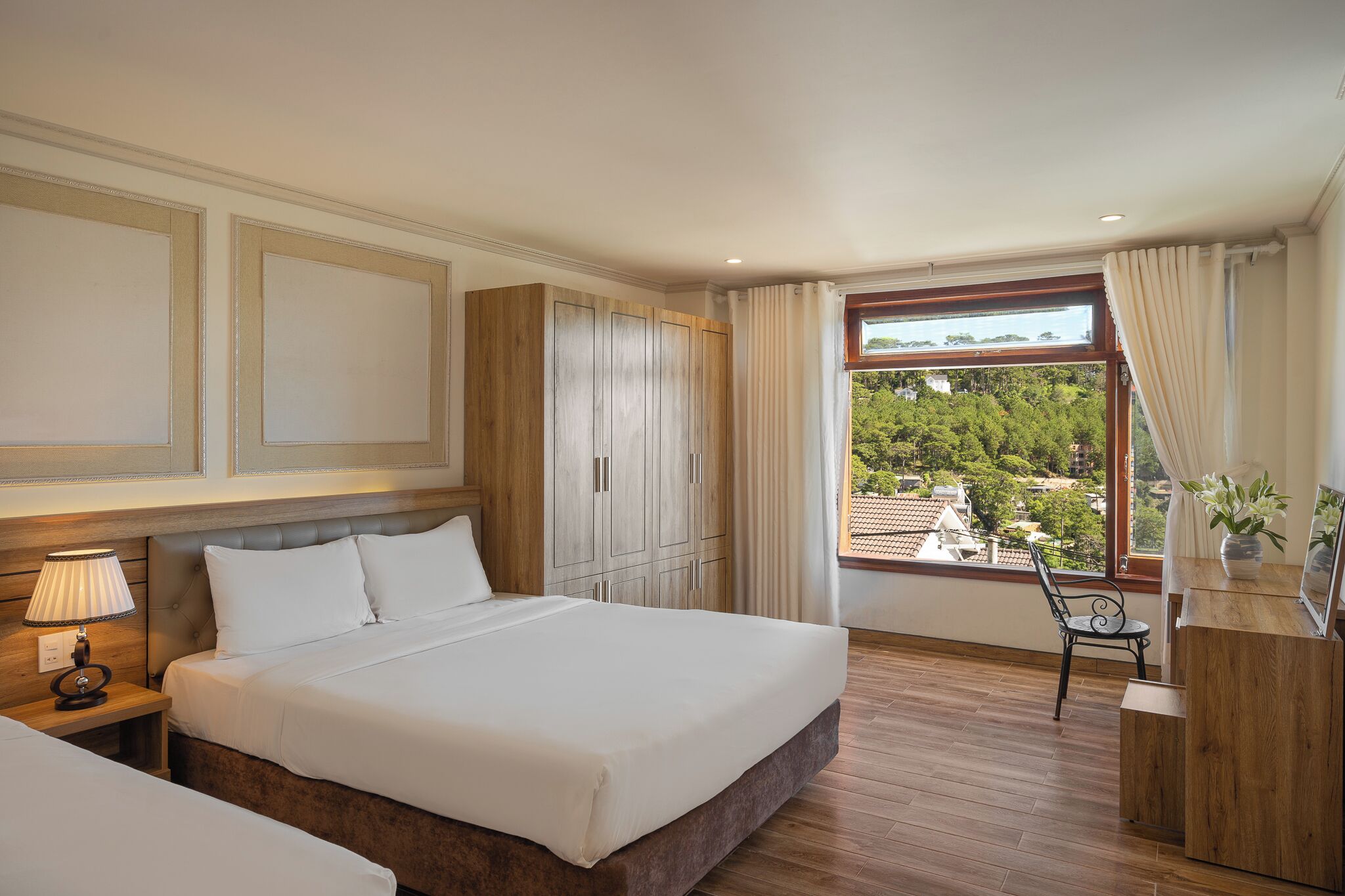 A hotel room with a large bed, a wooden wardrobe, a desk with a chair, and a window with a view of buildings and trees.