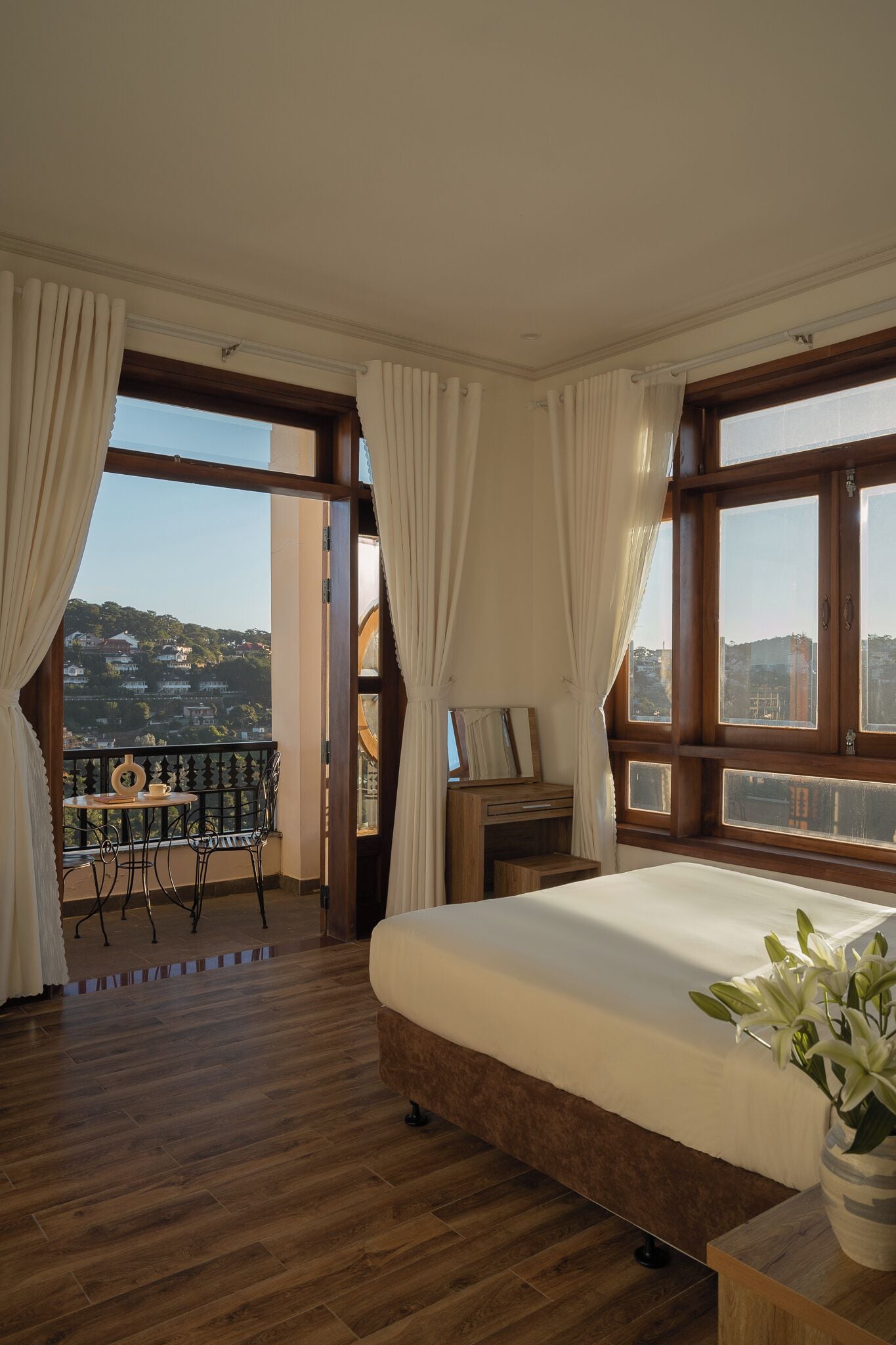 A bedroom with a large bed, wooden flooring, a balcony with a table and chairs, and a view of a hillside.