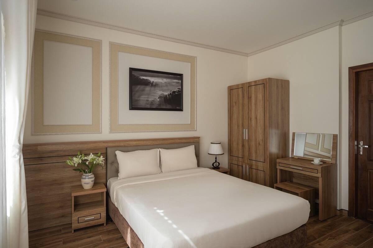 A bedroom with a large bed, wooden headboard, a nightstand with a lamp, a wardrobe, and a framed picture on the wall.