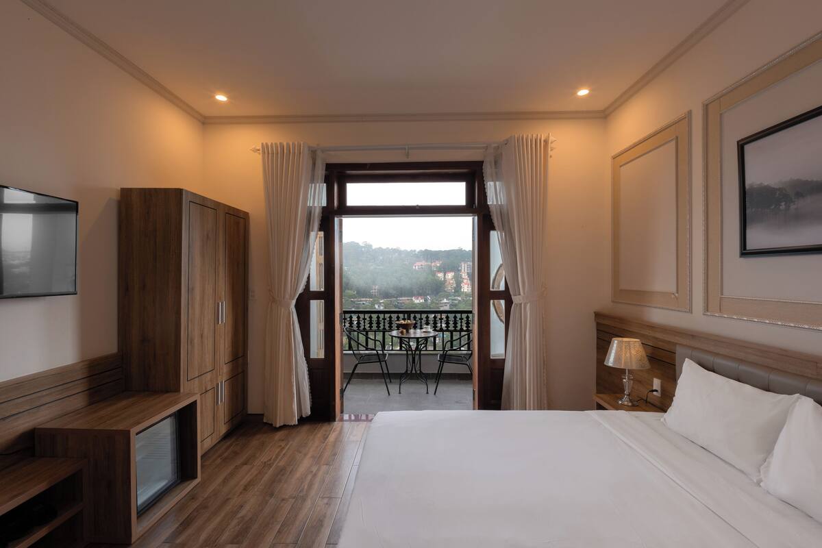 A hotel room with a large bed, a TV, wooden furniture, a balcony with a table and chairs, and a view of a cityscape.