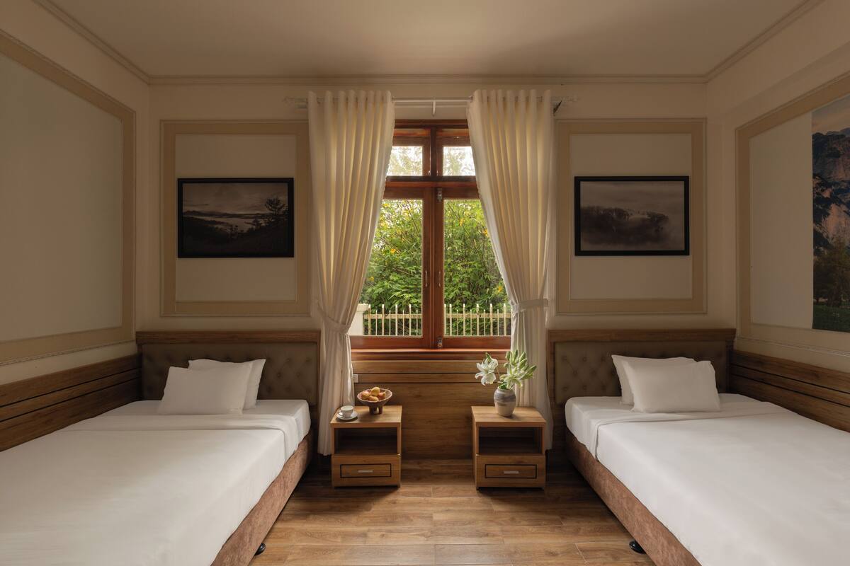 A room with two beds, a window with curtains, a small table with a bowl of fruit, and framed pictures on the walls.