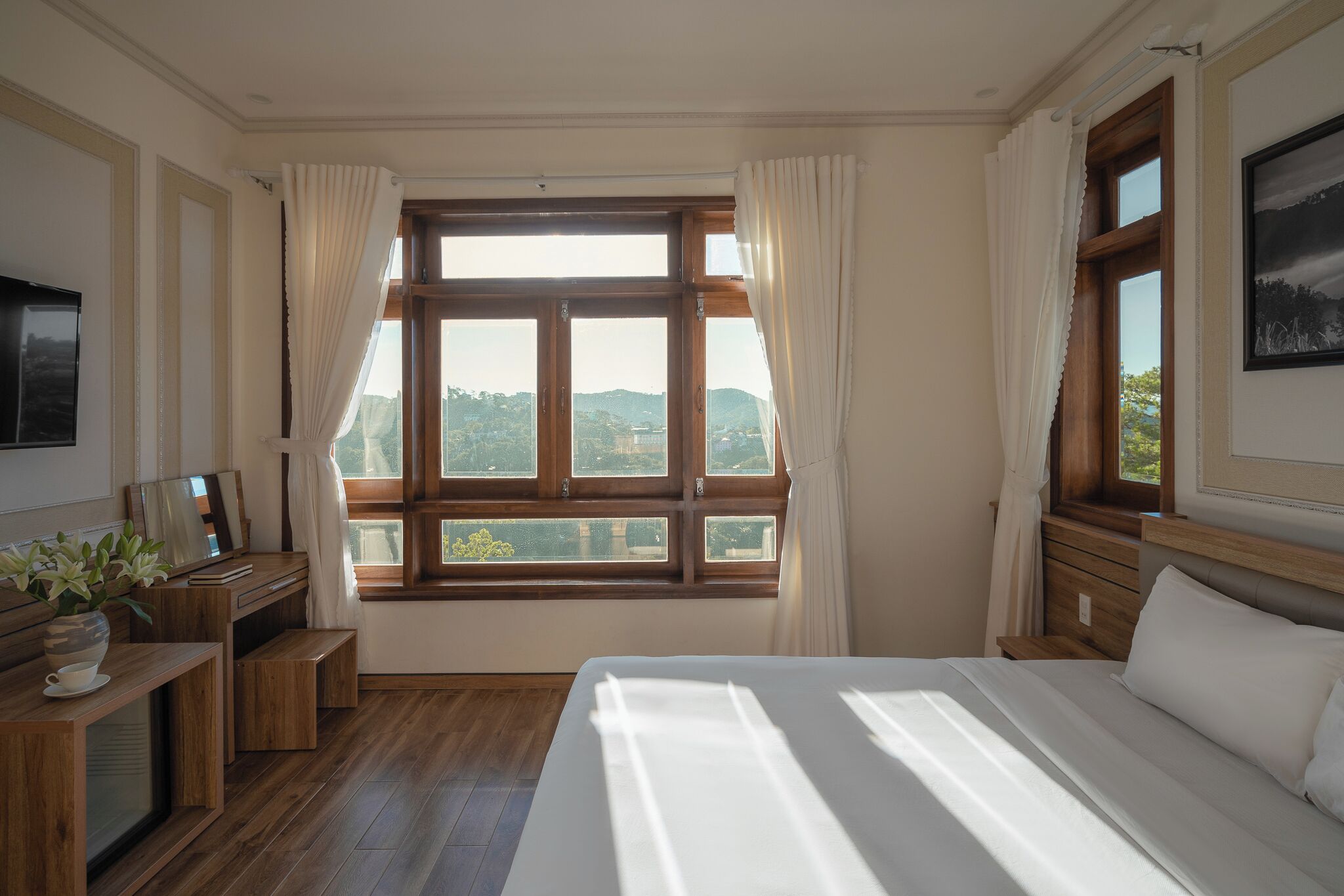 A bedroom with a large window, wooden furniture, and a bed with white bedding.