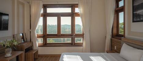 A bedroom with a large window, wooden furniture, and a bed with white bedding.