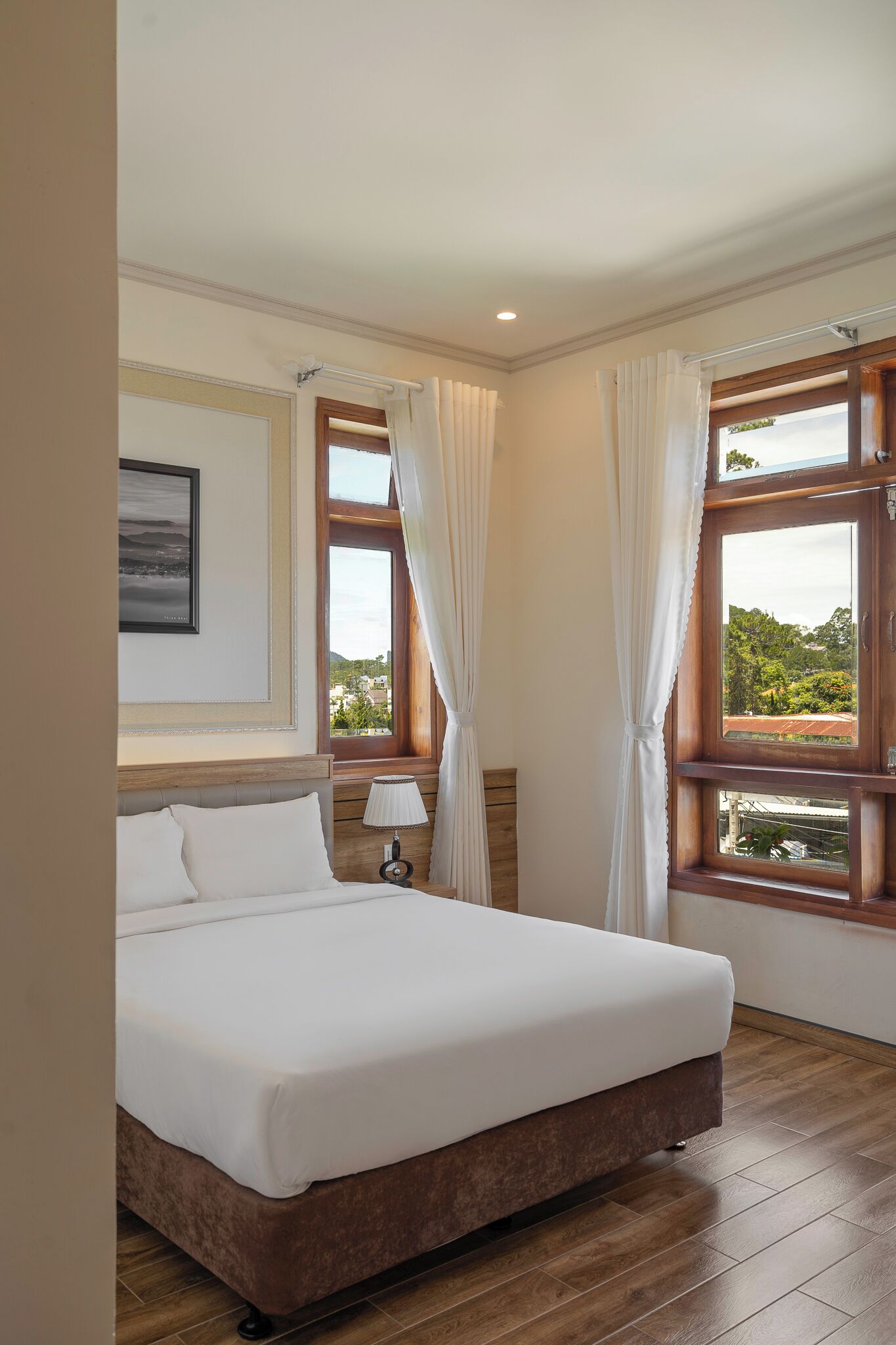 Ein Hotelzimmer mit einem großen Bett, zwei Vorhängen und Fenstern, einer Nachttischlampe und einem gerahmten Bild an der Wand.
