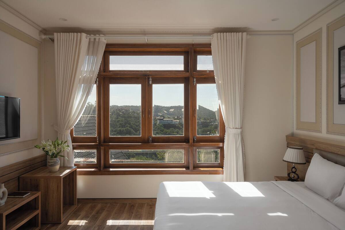 A hotel room with a large window, wooden furniture, a bed, a bedside table with a lamp, and a TV.